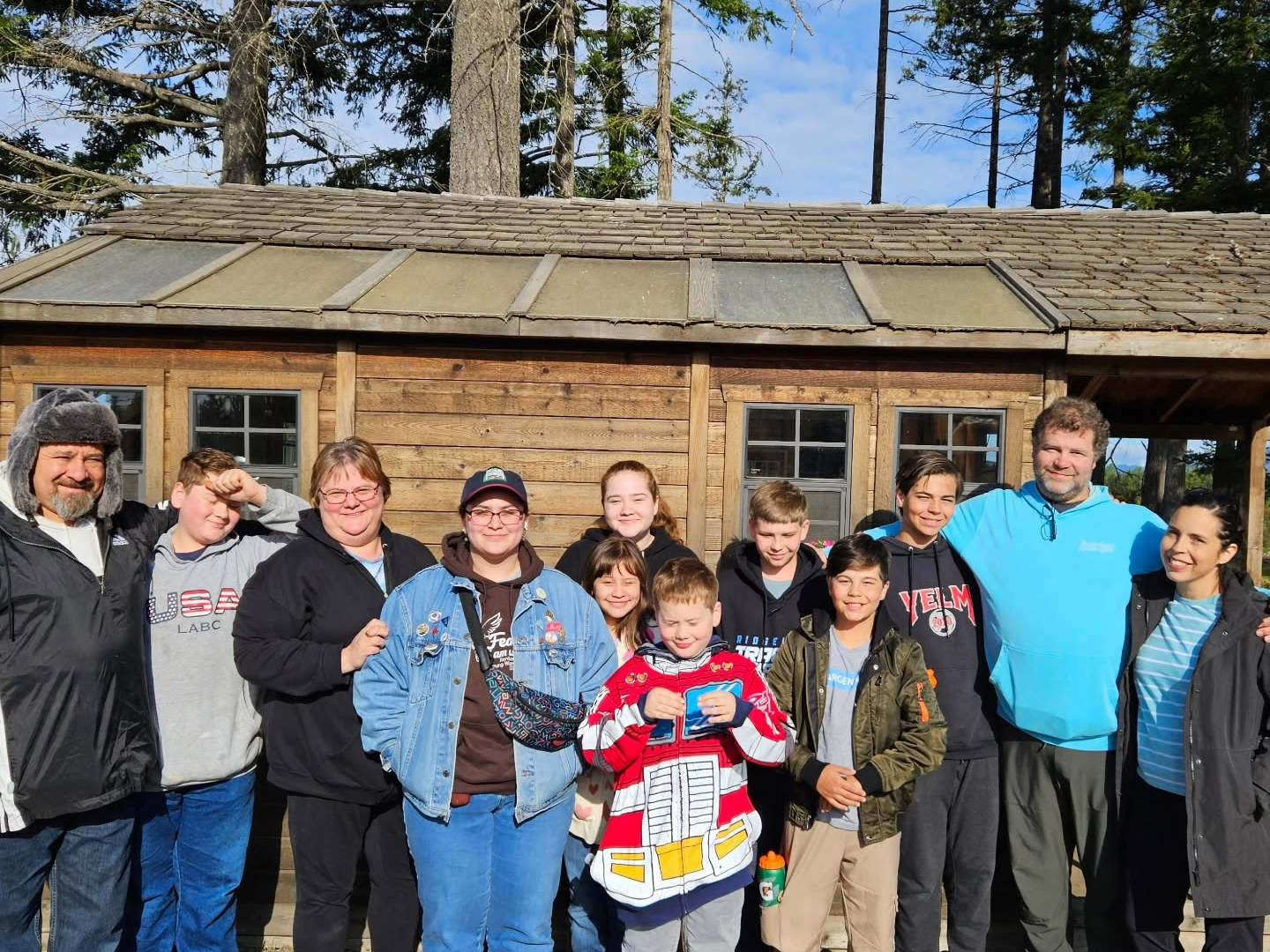 Group of people standing outside a wooden cabin with trees and blue sky in the background.