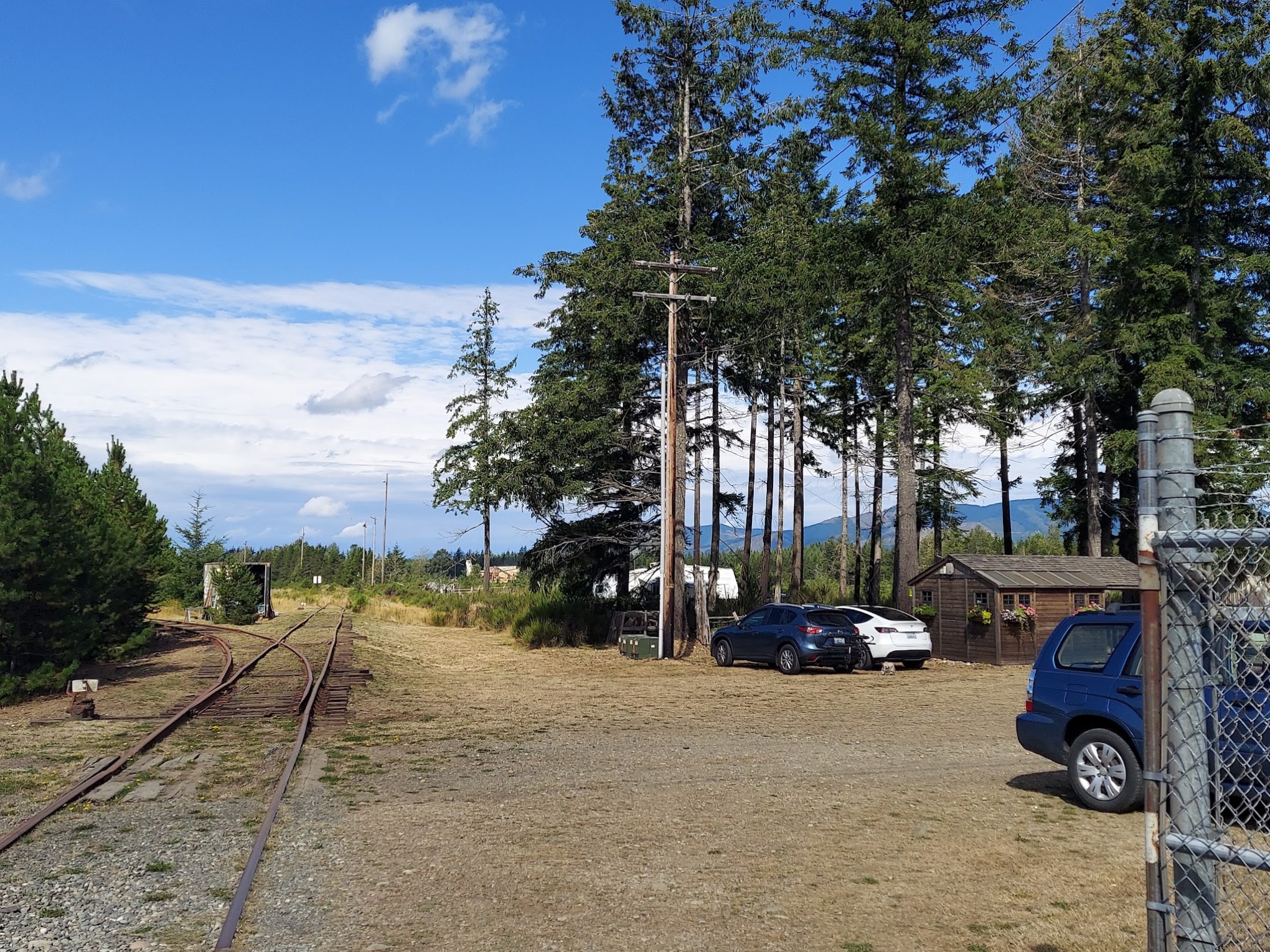 Rural area with railway tracks, cars, trees, and a fence under a blue sky.